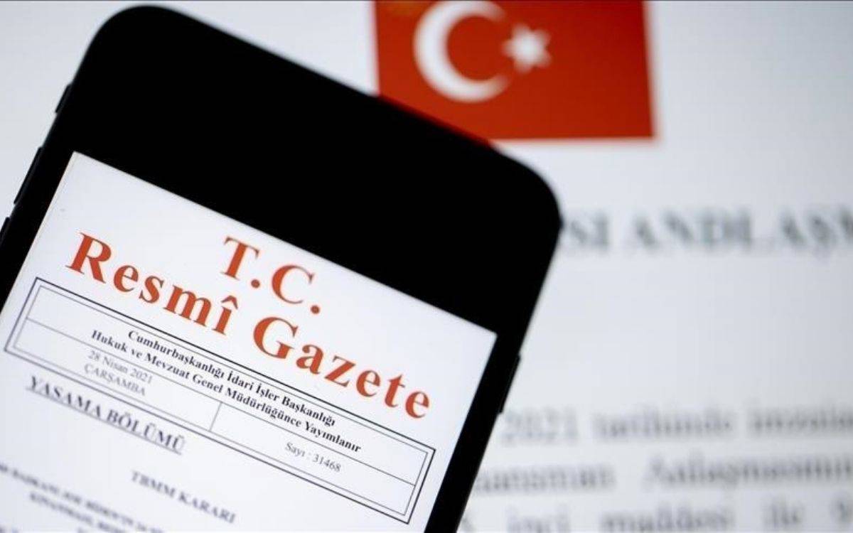 Bu görselde bir akıllı telefon ekranında T.C. Resmî Gazete ifadesi yer alıyor. Ekranda, Cumhurbaşkanlığı İdari İşler Başkanlığı'nın yayımladığı bir kararın üst kısmı görülüyor. Telefon ekranının arka planında ise Türk bayrağı ve üzerinde yazılar bulunan bir sayfa dikkat çekiyor. Resmî Gazete'nin klasik kırmızı renkli başlığı belirgin biçimde öne çıkıyor.