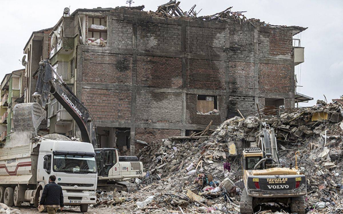 Fotoğrafta bir deprem sonrası yıkılmış bir binanın enkazı görülüyor. Yarı yıkılmış olan yapı, ağır hasarlı durumda ve üst katlarının bir kısmı çökmüş. Binanın önünde enkaz kaldırma çalışmaları devam etmekte. Bir iş makinesi (ekskavatör) enkazı bir kamyonun kasasına yüklerken başka bir iş makinesi de yıkıntılar arasında duruyor. Çevrede çeşitli moloz yığınları, kırık beton parçaları, demir çubuklar ve dağılmış eşyalar dikkat çekiyor. Yerde biriken enkaz miktarı oldukça fazla.