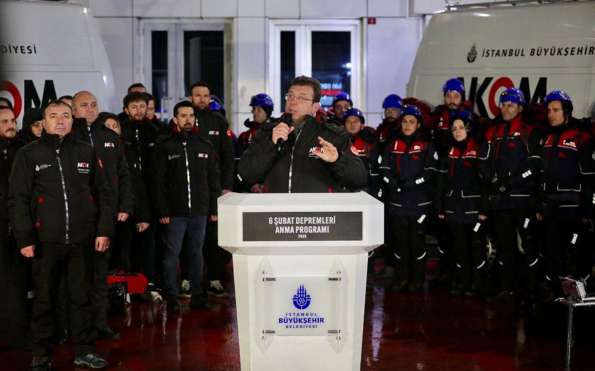 Fotoğrafta, İstanbul Büyükşehir Belediye (İBB) Başkanı Ekrem İmamoğlu, 6 Şubat Depremleri Anma Programı'nda konuşma yapıyor. İmamoğlu, AKOM (Afet Koordinasyon Merkezi) yazılı bir platformun arkasında, elinde mikrofonla katılımcılara hitap ediyor. Sahnenin arka tarafında AKOM'un kurtarma ekibi olduğu anlaşılan, kışlık giysiler ve güvenlik ekipmanları giymiş bir grup görevli yer alıyor. Çalışanlar montlarında İBB ve AKOM logolarını taşıyorlar.