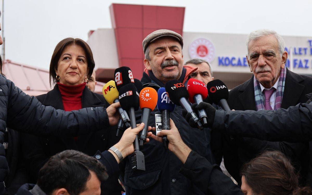 Fotoğrafta, basın açıklaması yapan bir grup siyasetçi ve onların etrafını saran gazeteciler görülüyor. Ortada, bir erkek siyasetçi mikrofonlara konuşuyor. Sağında kırmızı ceketli bir kadın ve solunda yaşlı bir erkek yer alıyor. Mikrofonlarda çeşitli medya kuruluşlarının logoları dikkat çekiyor. Arka planda bir kamu binasının tabelası ve kırmızı bir yapı görünmekte. Gazeteciler, mikrofonları uzatarak açıklamayı kaydediyor. Bu görüntü, önemli bir siyasi açıklama veya gelişmeyle ilgili bir basın toplantısını yansıtıyor.