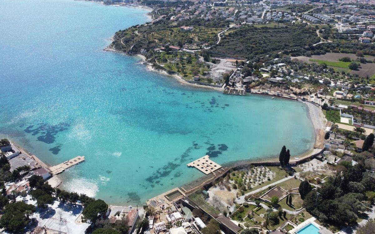 Fotoğraf, İzmir’in Çeşme ilçesinde bulunan dünyaca ünlü Aya Yorgi Koyu’nun havadan çekilmiş bir görüntüsünü gösteriyor. Turkuaz ve berrak sularıyla dikkat çeken koy, yarımay şeklinde bir kıyı hattına sahip. Deniz, kıyıya doğru giderek sığlaşıyor ve kumsalla buluşuyor.