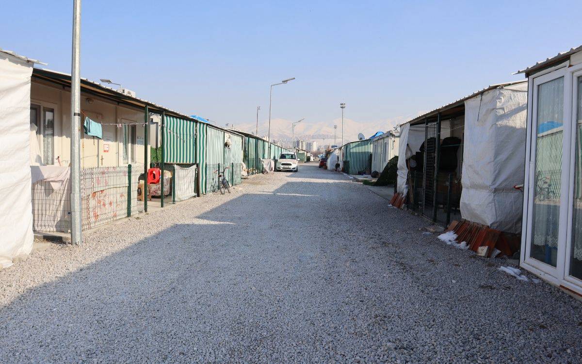 Fotoğrafta, bir konteyner kentten bir görüntü yer alıyor. Geniş, taş kaplı bir yolun iki yanında sıralanmış konteyner evler bulunuyor. Konteynerlerin bazıları brandalar ve tente örtülerle kaplanarak daha yaşanabilir hale getirilmiş. Kimi konteynerlerin önünde çamaşırlar asılı, bazıları ise tel çitlerle çevrelenmiş.