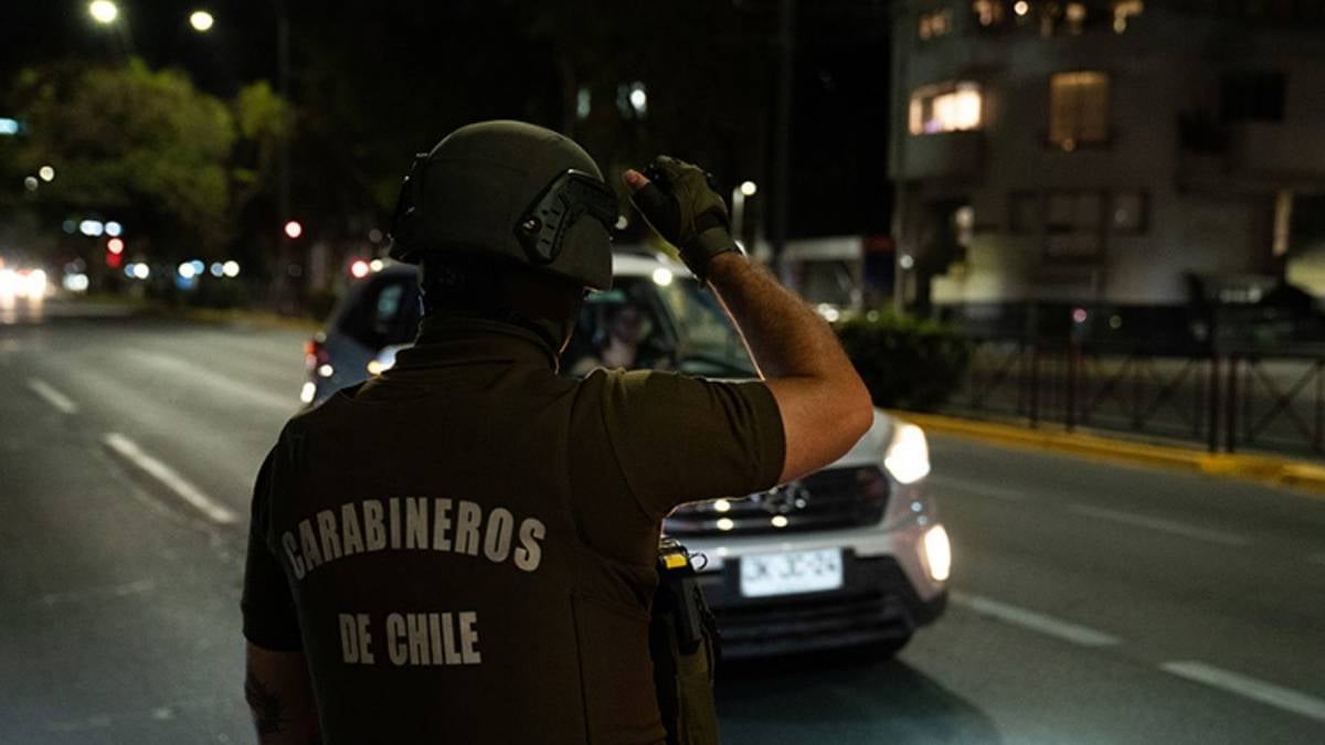 Bu fotoğraf gece saatlerinde Şili'de çekilmiş gibi görünüyor. Ön planda, üzerinde "Carabineros de Chile" yazan koyu yeşil üniformasıyla bir güvenlik görevlisi (muhtemelen Şili polisi veya askeri personel) bulunuyor. Kafasında koruyucu bir kask var ve sağ elini kaldırarak bir aracı durduruyor.