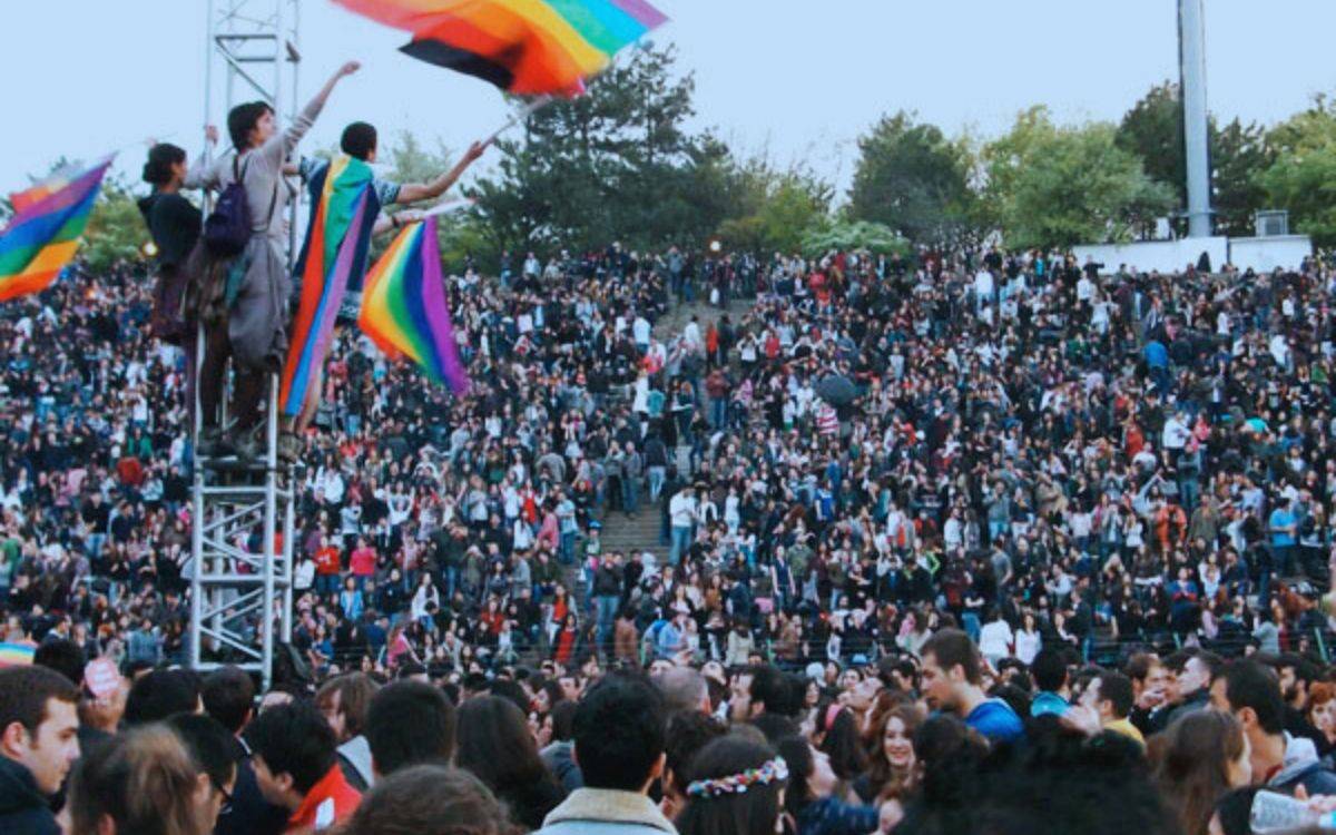 Fotoğrafta, büyük bir açık hava alanında kalabalık bir insan topluluğu görülüyor. Çok sayıda kişi, bir amfi tiyatro ya da geniş bir merdivenli alanda toplanmış, coşkulu bir atmosfer içinde etkinliğe katılıyor.  Ön planda, gökkuşağı bayrakları taşıyan birkaç kişi bir metal yapının üzerine çıkmış ve bayrakları dalgalandırıyor. Ellerini havaya kaldırmış olan bu kişiler, coşkulu bir şekilde kalabalığa sesleniyor ya da etkinliğin bir parçası olarak heyecanlarını paylaşıyor.