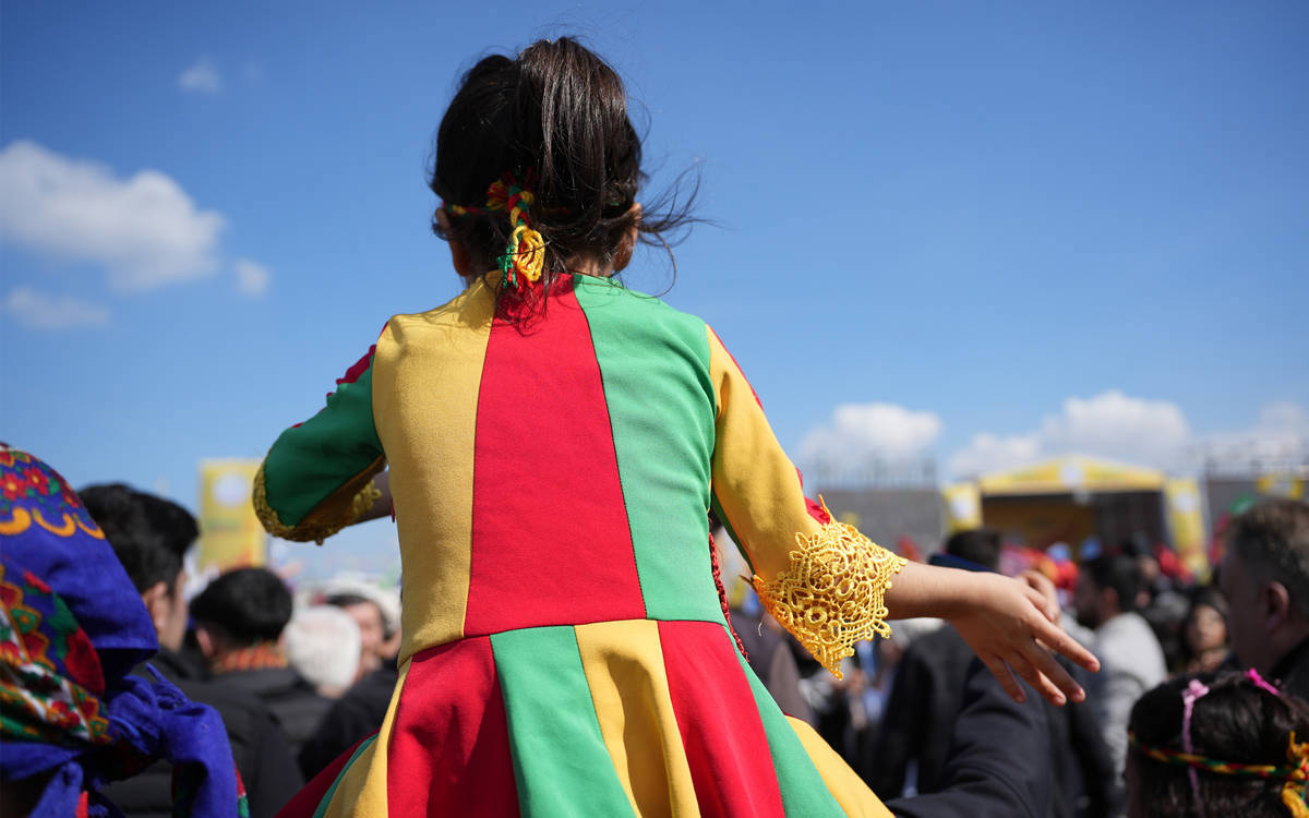 Fotoğrafta sarı kırmız yeşil renklerde elbise giymiş bir çocuk var. Çocuğun Newroz kutlamasında olduğu anlaşılıyor.