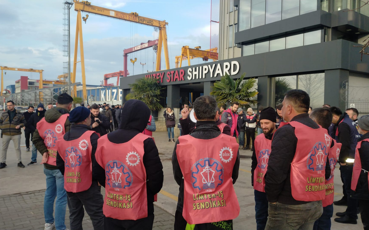 Fotoğrafta bir grup işçi, Kuzey Star Shipyard (tersanesi) yazısının bulunduğu modern bir bina önünde toplanmış durumda. Çoğu kişi pembe-kırmızı renkte yelekler giyiyor ve bu yeleklerin arkasında LİMTER-İŞ SENDİKASI ve DİSK logoları bulunuyor.