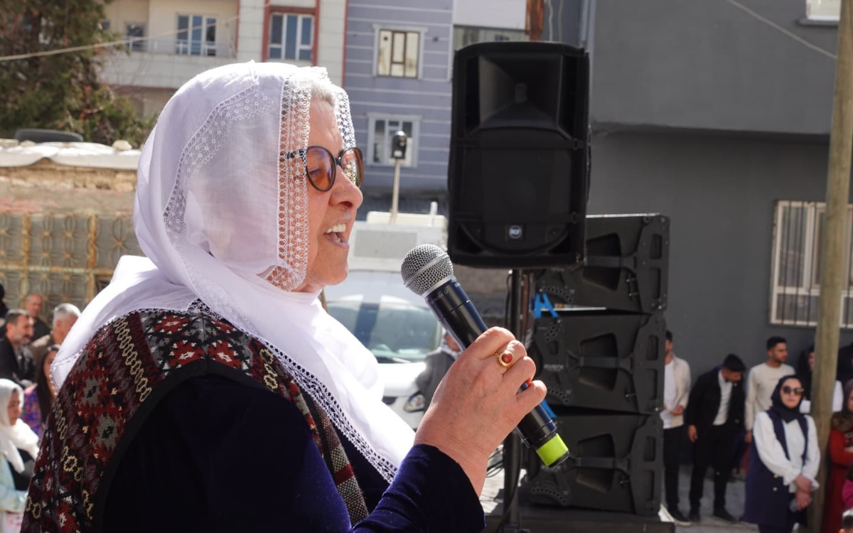 Fotoğrafta, Barış Annesi Havva Kıran var. Kıran, elinde mikrofonla konuşma yapıyor. Beyaz, işlemeli bir başörtüsü takmış ve üzerine siyah, kadife benzeri uzun kollu bir kıyafet giymiş. Kıyafetinin üzerine renkli desenleri olan bir yelek giymiş. Yüzünde kahverengi çerçeveli gözlük var ve parmağında büyük, taşlı bir yüzük bulunuyor. Kıran, açık havada, bir etkinlikte konuşma yapıyor gibi görünüyor. Arka planda bir grup insan onu dinliyor; bazı kadınlar beyaz başörtüsü takarken, erkekler takım elbise veya gündelik kıyafetler giymiş. Büyük siyah hoparlörler dikkat çekiyor, bu da bir miting, anma töreni veya topluluk etkinliği olduğunu düşündürüyor. Binalar, elektrik direkleri ve arabalar, etkinliğin şehir içinde bir mahallede gerçekleştiğini gösteriyor. Hava açık ve güneşli, ışık doğrudan yüzlere vuruyor.