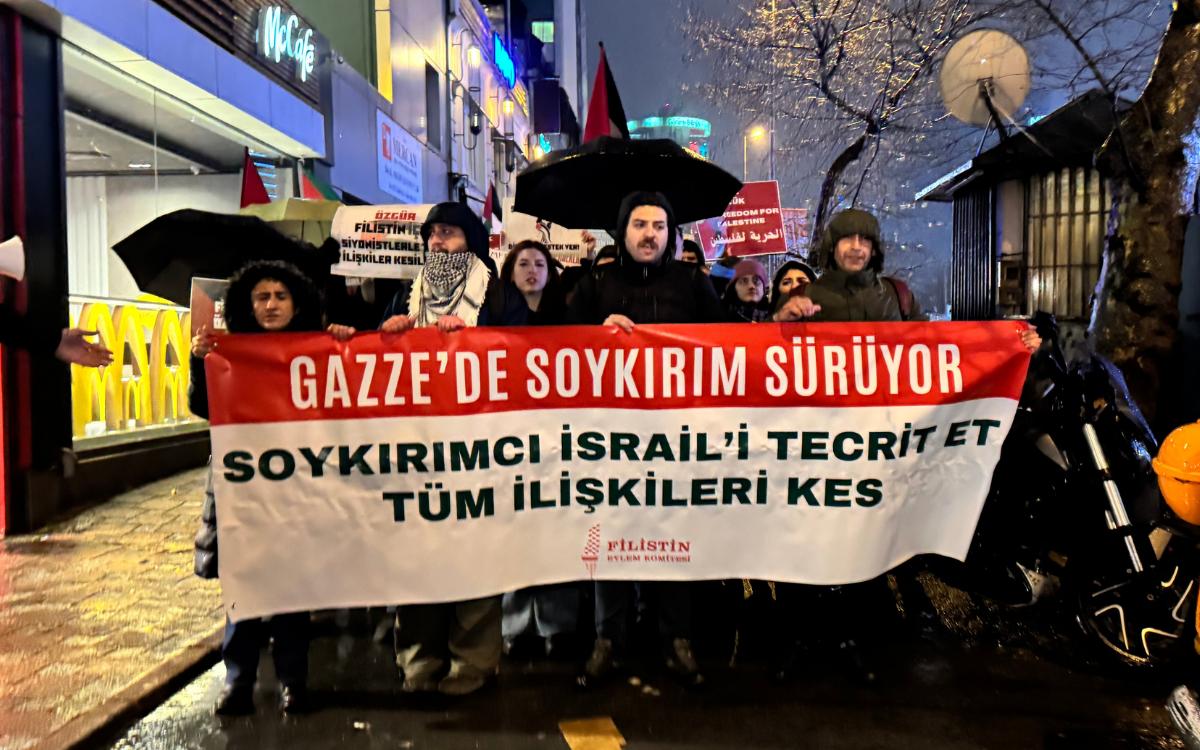 Fotoğrafta kitle “Gazze’de soykırım sürüyor. Soykırımcı İsrail’i tecrit et. Tüm ilişkileri kes” yazılı pankart ile yürüyor. Arkada Filistin bayrakları ve şemsiyeler gözüküyor.