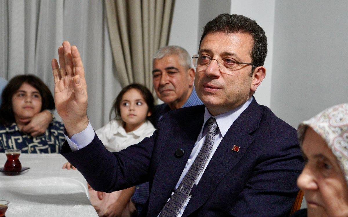 Bu fotoğrafta, takım elbise giymiş bir erkek (muhtemelen bir politikacı) bir masa etrafında oturmuş, sağ elini havaya kaldırmış bir şekilde konuşuyor ya da bir konuya dikkat çekiyor gibi görünüyor. Üzerinde küçük bir Türk bayrağı rozeti ve mikrofon iğnesi bulunuyor.