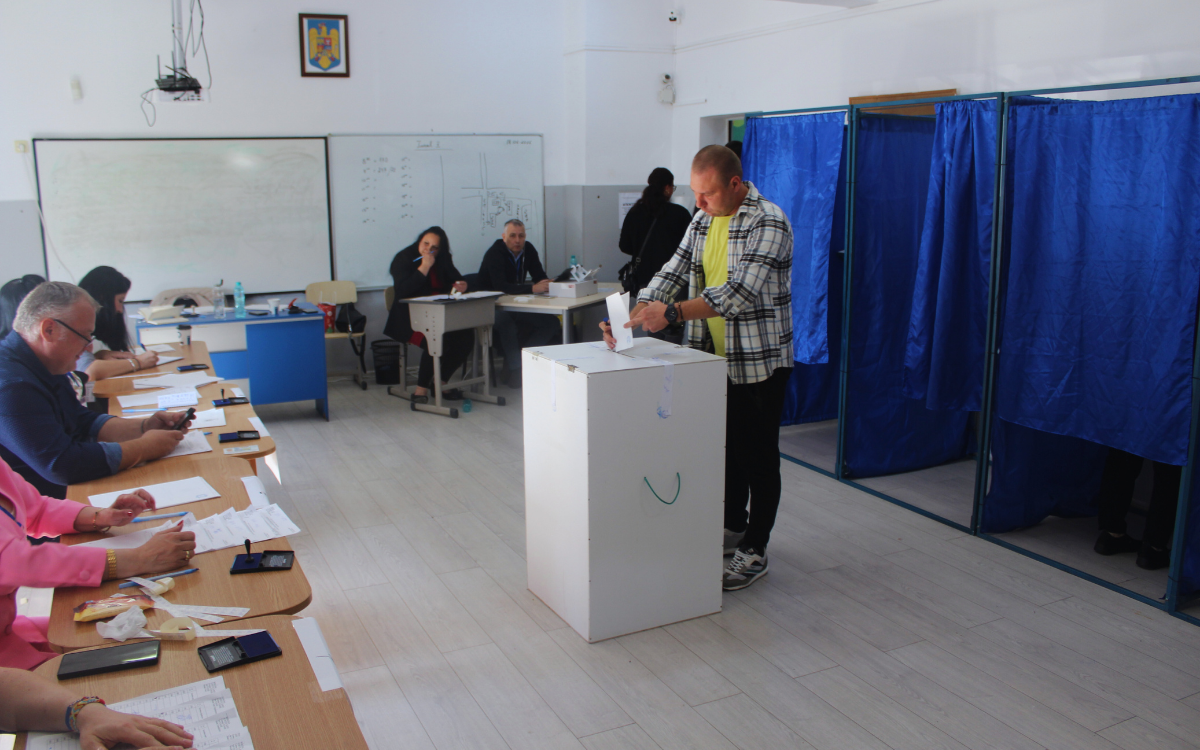 Fotoğrafta bir kişi sandığa oy atıyor etrafında masalarda oturan gözetmenler var.