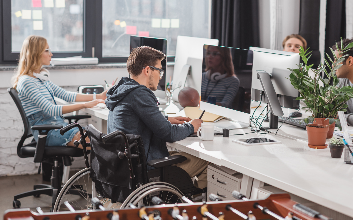 Bir ofis ortamında dört kişi bilgisayar başında çalışıyor. Ön planda tekerlekli sandalye kullanan bir erkek, masa başında bilgisayar ekranına bakıyor. Yanında bir kahve kupası, defter ve kalem var. Sol arkada, kulaklık takan sarı saçlı bir kadın çalışıyor. Masada saksı bitkileri, tablet ve çeşitli ofis eşyaları bulunuyor. Arka planda pencere, siyah perdeler ve yapışkan notlar görülüyor.