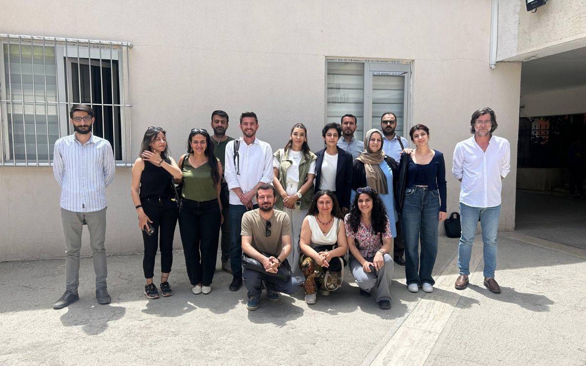 Fotoğrafta, bir grup insan açık havada, bir binanın önünde topluca poz veriyor. Görüntüde 17 kişi yer alıyor; 12’si ayakta duruyor, 5’i önde çömelmiş ya da oturur pozisyonda.