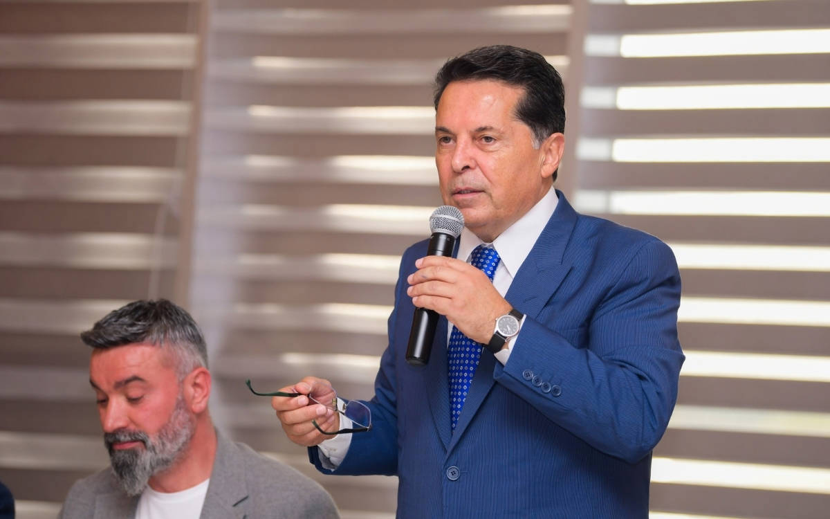 Fotoğrafta iki erkek görülüyor. Ön planda, mavi çizgili bir takım elbise ve mavi-beyaz puanlı kravat giymiş bir adam, sağ elinde mikrofon, sol elinde gözlükle konuşma yapıyor. Ciddi ve kararlı bir ifade ile konuşmasını sürdürüyor. Bileğinde metal kayışlı bir saat takılı. Koyu renk saçları düzgün taranmış. Arka planda yer alan yatay çizgili stor perdeler mekânın bir iç mekan toplantı salonu olduğunu düşündürüyor.  Ön plandaki kişinin solunda yer alan başka bir erkek ise gri ceket ve beyaz tişört giymiş, başını hafifçe öne eğmiş, gözlerini kapatmış ya da aşağı bakıyor. Saçları ve sakalı kırlaşmış ve oldukça belirgin. Bu kişinin duruşu daha sakin ve sessiz bir izlenim veriyor.