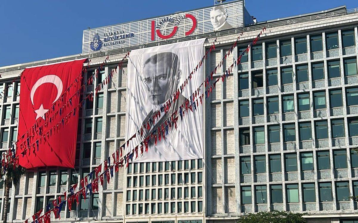 Bu fotoğraf, İstanbul Büyükşehir Belediyesi’nin (İBB) Saraçhane’deki ana hizmet binasını güneşli bir günde cepheden göstermektedir. Bina ve Süslemeler: Binanın üst kısmında “İstanbul Büyükşehir Belediyesi” yazısı ve İBB’nin logosu yer almaktadır. Hemen üstte, “Türkiye Cumhuriyeti’nin 100. yılı”na atıfla kullanılan büyük kırmızı "100" rakamı ve yanında Atatürk portresi yer almaktadır. Binanın ön cephesine iki büyük afiş asılmıştır: Solda, kırmızı zemin üzerine beyaz ay-yıldızdan oluşan büyük Türk bayrağı yer almakta. Ortada ise Mustafa Kemal Atatürk’ün siyah-beyaz büyük bir portresi bulunmaktadır. Bu iki afişin üzerine çapraz şekilde çekilmiş olan kırmızı ve mavi üçgen flamalar, coşkulu bir kutlama atmosferi oluşturmaktadır. Flamaların üzerinde de ay-yıldız sembolleri görülmektedir.