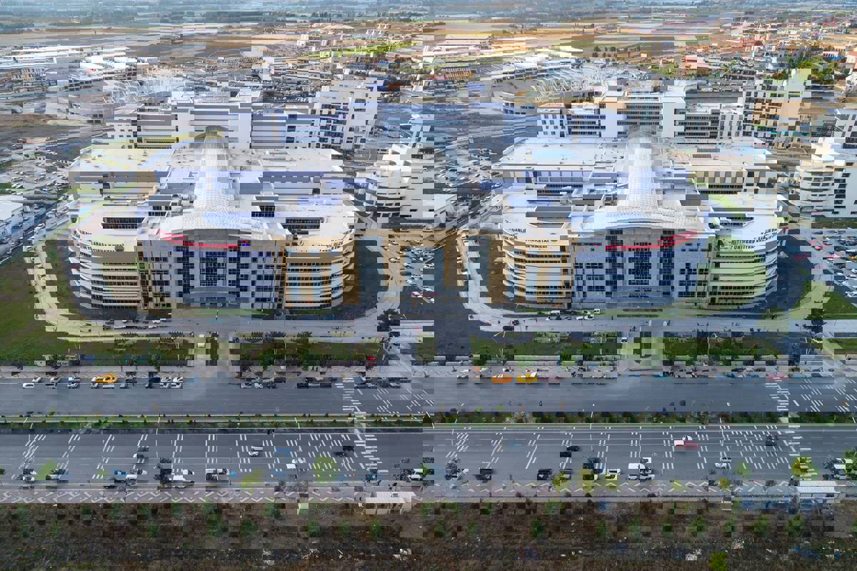 Fotoğrafta, modern ve büyük ölçekli bir hastane kompleksi yukarıdan (kuşbakışı) görüntülenmiş. Binanın ön cephesi geniş camlarla kaplı ve sarı-bej tonlarında taş cepheyle modern bir mimari tarzı yansıtıyor. Üzerinde “T.C. Sağlık Bakanlığı – MÜTF Üniversitesi Eğitim ve Araştırma Hastanesi” yazısı net şekilde görülüyor. Binanın sol ve sağ kanatları kavisli bir yapıya sahip ve ortada büyük bir ana giriş yer alıyor. Girişin önünde araçların giriş-çıkış yaptığı bir yol ve hastane bahçesi dikkat çekiyor.  Hastanenin çevresinde geniş yollar, park alanları ve başka binalar yer alıyor. Arka planda ise açık tarım arazileri ve çeşitli yapılaşmalar görünmekte. Ön planda, çift yönlü trafiğe açık geniş bir cadde ve yol kenarına park etmiş birçok araç seçilebiliyor.