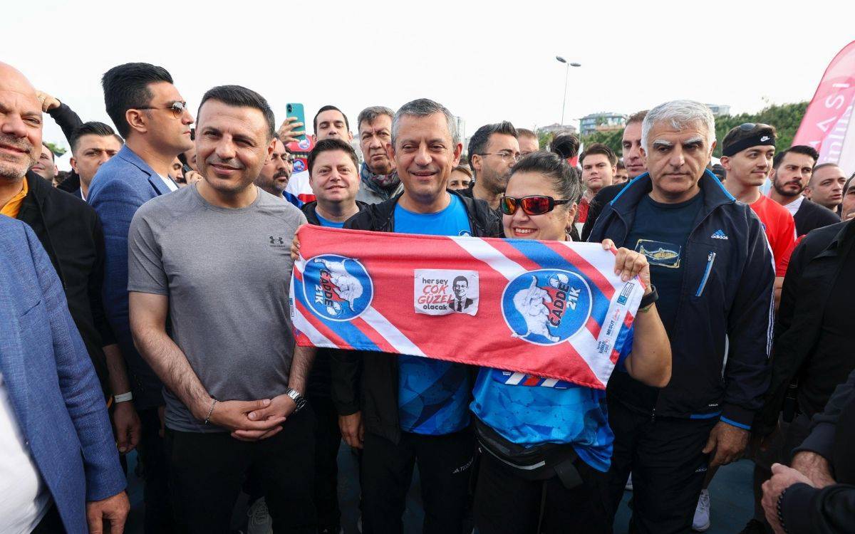 Fotoğrafta, kalabalık bir grup insan bir araya gelmiş ve gülümseyerek poz veriyor. Fotoğrafın merkezinde, CHP Genel Başkanı Özgür Özel yer alıyor. Spor kıyafetleri giymiş olan Özel’in sol yanında Kadıköy Belediye Başkanı Mesut Kösedağı bulunuyor. Sağ tarafta ise kırmızı gözlük takmış bir kadın, elinde “Cadde 21K” yazılı ve üzerinde Ekrem İmamoğlu’nun “Her şey çok güzel olacak” sloganının yer aldığı bir atkı tutuyor.  Atkıda ayrıca koşunun logosu ve organizasyon bilgileri de yer almakta. Arkada ise etkinliğe katılan çok sayıda kişi bulunuyor; bazıları cep telefonlarıyla fotoğraf çekiyor. Katılımcıların çoğu spor kıyafetleriyle etkinliğe hazır görünmekte. Genel atmosfer enerjik, birlik ve dayanışma duygusunu yansıtan bir kitle görüntüsü sunuyor. Etkinliğin bir koşu organizasyonu olduğu ve politik bir mesaj da içerdiği net biçimde görülüyor.