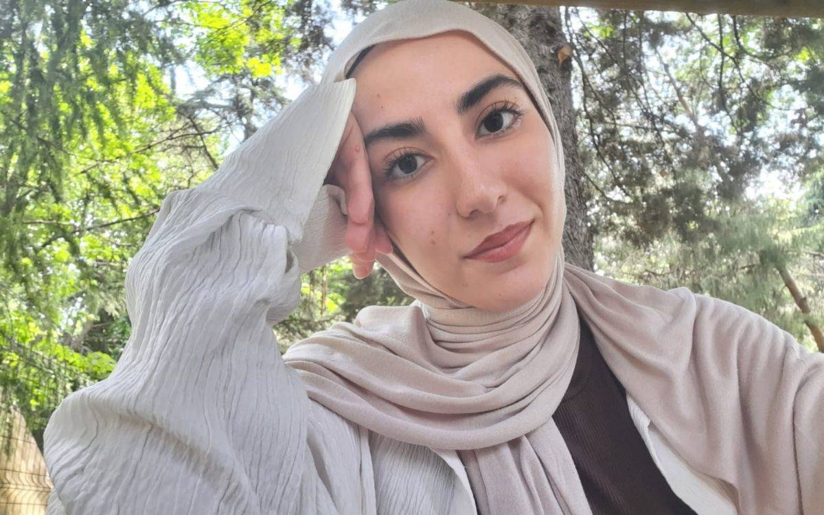 Fotoğrafta genç bir kadın, doğal ışıkla aydınlanmış yeşillikler içinde bir arka planda görülüyor. Kadın, başörtülü ve açık bej tonlarında bir şal takıyor. Üzerinde, zarif dokulu, hafif pileli ve bol kesimli beyaz bir bluz bulunuyor. Sağ elini yanağına dayamış, yüzünü hafifçe eğmiş bir şekilde kameraya bakıyor. Yüz ifadesi sakin, huzurlu ve dikkatle odaklanmış; dudaklarında belirsiz ama sıcak bir tebessüm var.