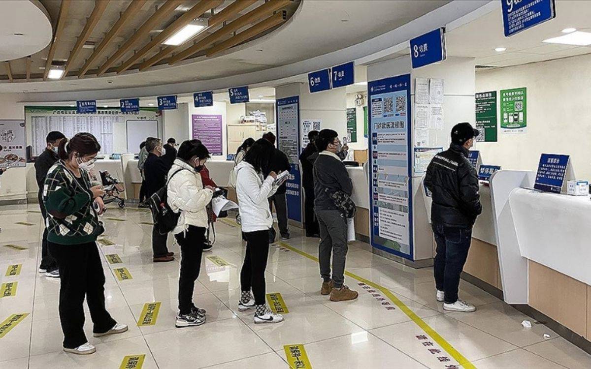 Fotoğrafta, çoğunluğu genç ve orta yaşlı bireylerden oluşan bir grup insanın, sıralarına uygun şekilde beklediği bir iç mekân görülüyor. Burası büyük olasılıkla bir hastanenin ya da sağlık merkezinin kayıt veya danışma bölümü. Zeminde sarı renkli şeritler ve üzerine Çince yazılar olan sosyal mesafe etiketleri dikkat çekiyor; bu da salgınla mücadele kapsamında alınan önlemleri yansıtıyor.  İnsanların çoğu maske takmış durumda ve ellerinde evraklar ya da telefonlar taşıyorlar. Sıra bekleyenler arasında beyaz montlu, siyah pantolonlu bir kadın ve onun hemen arkasında yeşil desenli kazak giymiş bir başka kadın göze çarpıyor. Her iki kişi de cep telefonlarına odaklanmış durumda. Danışma bankoları numaralandırılmış – 1’den 9’a kadar mavi tabelalarla gösterilmiş. Her bankonun üzerinde işlem türlerine dair açıklamalar yer alıyor.