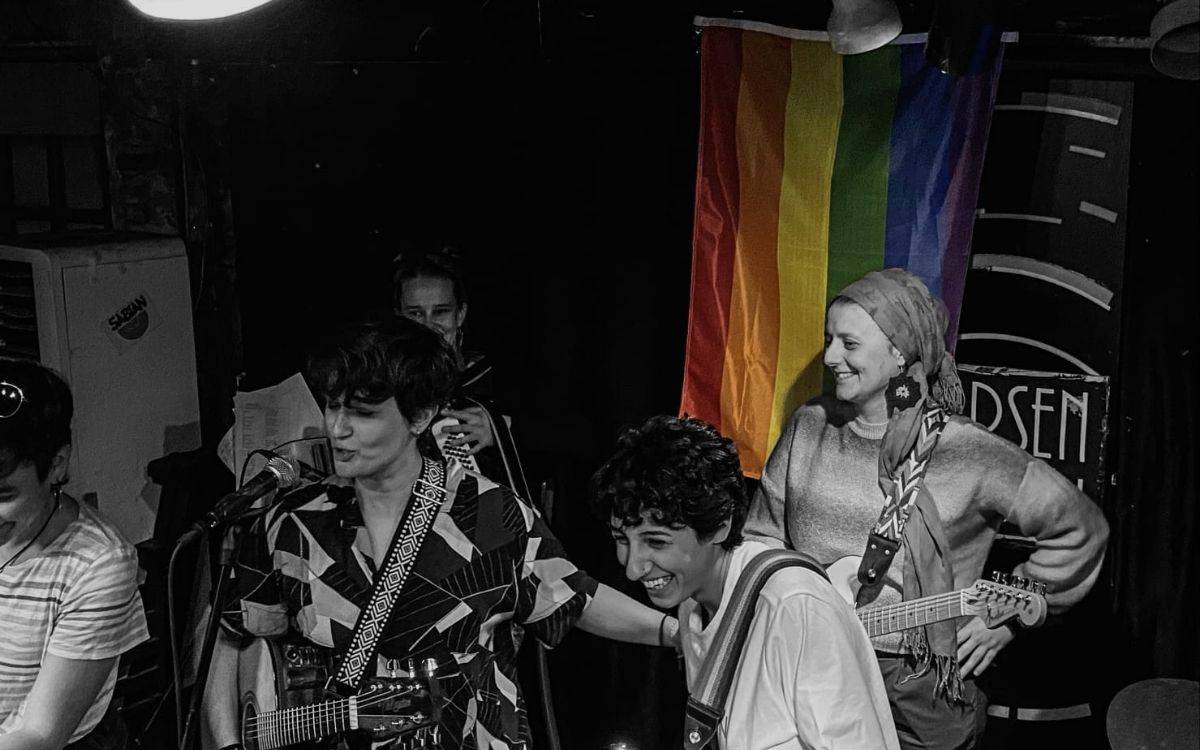 Fotoğrafta, sahnede birlikte müzik yapan ve keyifli anlar paylaşan bir grup insan yer alıyor. Görselin odak noktasında gitar çalan ve mikrofon başında şarkı söyleyen kişiler bulunuyor. Sağda başörtülü bir kadın gitarıyla gülümsüyor. Sahnenin arkasında ise büyük bir gökkuşağı bayrağı (LGBTİ+ bayrağı) asılı, bu da mekânın kapsayıcı ve queer dostu bir alan olduğunu gösteriyor.