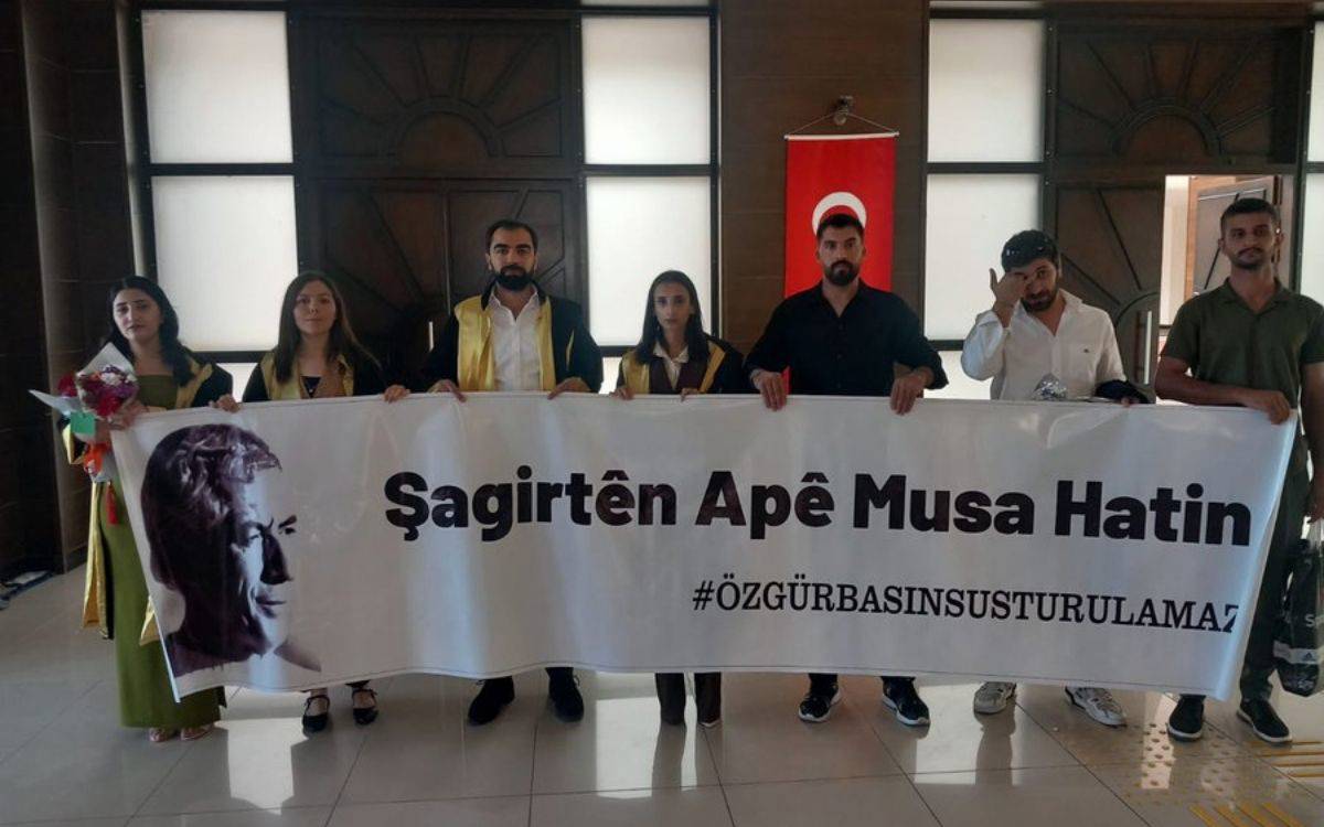 Bu fotoğraf, bir grup üniversite öğrencisinin mezuniyet töreni öncesi ya da sırasında birlikte tuttukları pankartla verdikleri toplumsal bir mesajı yansıtıyor.  Görselin detaylı betimlemesi: Ön planda, sekiz genç bir pankart tutuyor. Dördü mezuniyet cübbesi giymiş durumda; bu da onların yeni mezun olduklarını gösteriyor. Diğerleri sivil kıyafetli.  Gençlerin ellerinde tuttuğu beyaz pankartta büyük puntolarla Kürtçe “Şagirtên Apê Musa Hatin” (Apê Musa’nın öğrencileri geldi) yazıyor. Altında ise Türkçe bir hashtag yer almakta: #ÖzgürBasınSusturulamaz.