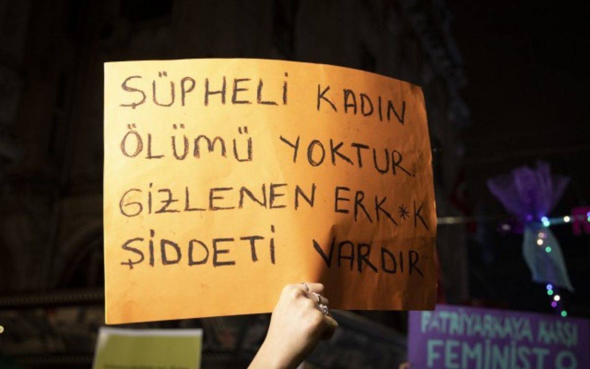 u fotoğrafta, gece saatlerinde bir protesto ya da eylem sırasında çekilmiş bir görüntü yer alıyor. Görselde, turuncu renkte bir kartona siyah keçeli kalemle yazılmış bir döviz tutuluyor. Dövizde şu ifadeler yer almakta: "ŞÜPHELİ KADIN ÖLÜMÜ YOKTUR. GİZLENEN ERK*K ŞİDDETİ VARDIR"