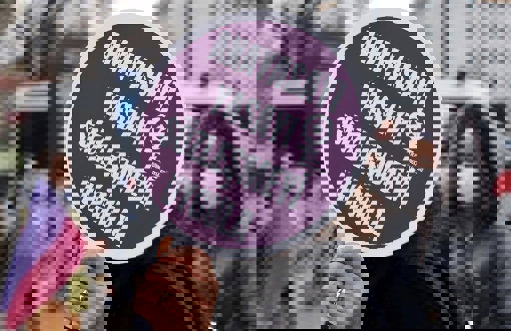 Bu fotoğraf bir protesto veya eylem sırasında çekilmiş. Görselin odak noktasında, bir kişi mor zeminli yuvarlak bir döviz tutuyor. Dövizde beyaz büyük harflerle yazılmış şu ifade yer alıyor:  “Anayasayı, yasayı, sözleşmeyi uygula”