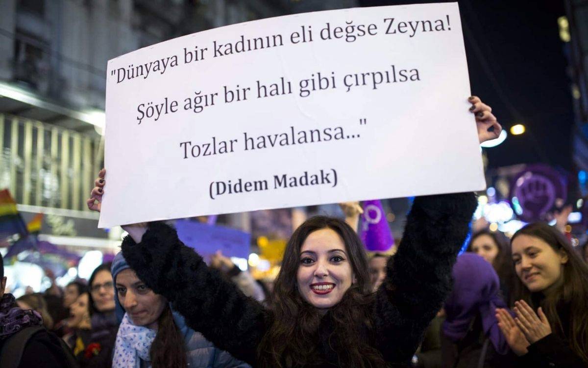 Fotoğrafta, gece saatlerinde bir şehir meydanında düzenlenen kalabalık bir kadın yürüyüşü veya mitinginden bir sahne yer alıyor. Ön planda, genç bir kadın yüzünde kararlı ve neşeli bir ifadeyle kamera objektifine bakarak elinde beyaz bir pankart tutuyor.