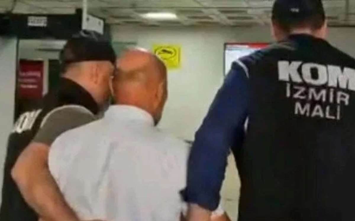 Bu fotoğrafta, İzmir Adalet Sarayı'nın dış cephesinden bir bölüm yer alıyor. Görselde büyük, altın sarısı renkte harflerle yazılmış şu ifadeler dikkat çekiyor:  "T.C. İZMİR ADALET SARAYI"  Yazılar, koyu renkli büyük taş bloklardan oluşan bir duvar üzerine monte edilmiş. Harflerin üç boyutlu olması ve gölgelerinin duvarda belirginleşmesi yazılara vurgu katıyor. Sağ alt köşede çiçek açmış kırmızı renkli küçük bir bitki de kadraja girmiş. Görsel, devlet kurumlarına özgü ciddi ve resmi bir atmosfer yansıtıyor.