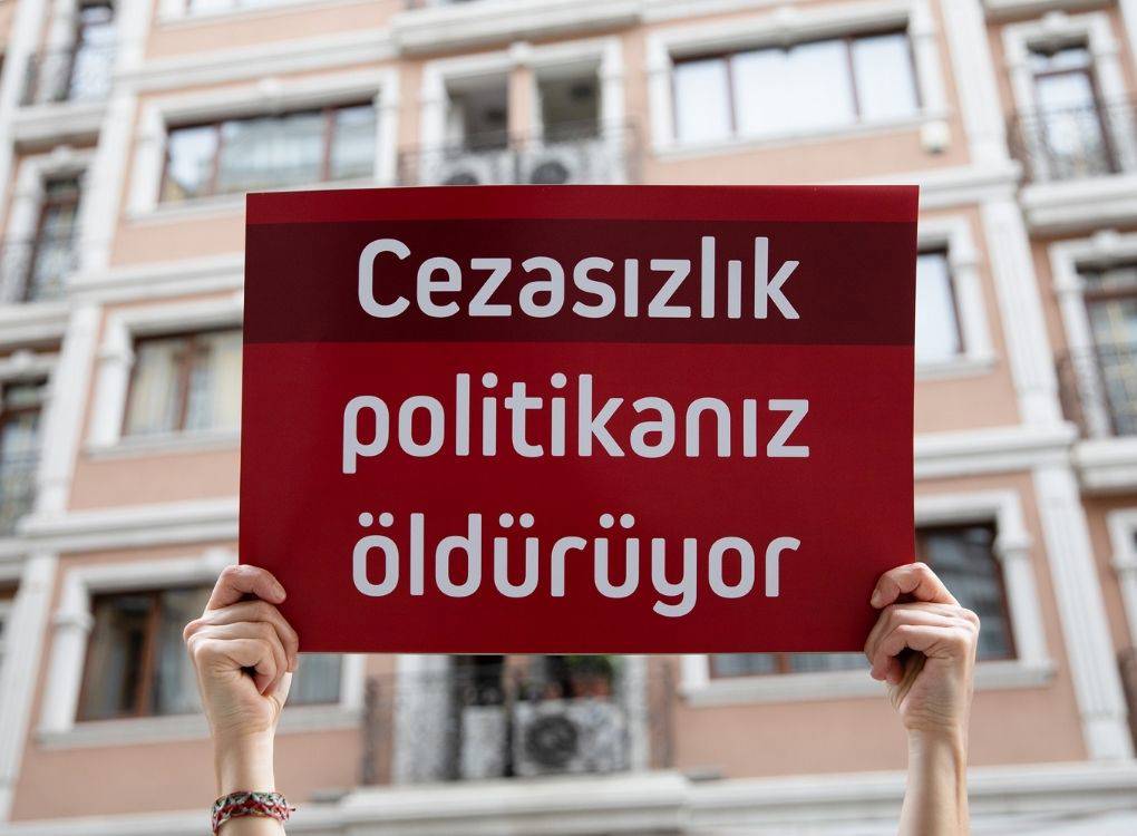 Bir kişinin iki eliyle tuttuğu kırmızı renkli bir protesto pankartı ön planda yer alıyor. Pankartta beyaz, kalın harflerle şu yazı dikkat çekiyor: “Ceza­sızlık politikanız öldürüyor” Yazı üç satıra yayılmış durumda ve güçlü bir mesaj içeriyor.