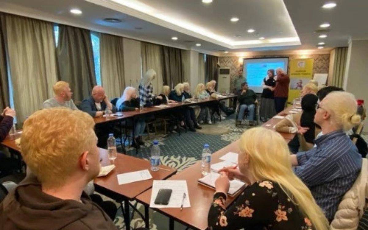 Fotoğrafta, dikdörtgen biçiminde düzenlenmiş uzun masaların etrafında oturan çok sayıda kişi görülüyor. Katılımcılar, önlerinde defter ve kalemlerle notlar alıyor, masalarda su şişeleri var. Oda, büyük pencerelerden giren doğal ışıkla aydınlanmış, pencerelerde uzun perdeler bulunuyor. Arka planda, bir projeksiyon perdesine yansıtılmış sunum ve yanında sarı renkli bir tanıtım panosu yer alıyor. Sunum yapan iki kişi önde ayakta duruyor, dinleyiciler dikkatle onları izliyor. Mekân, otel ya da toplantı salonu havasında.