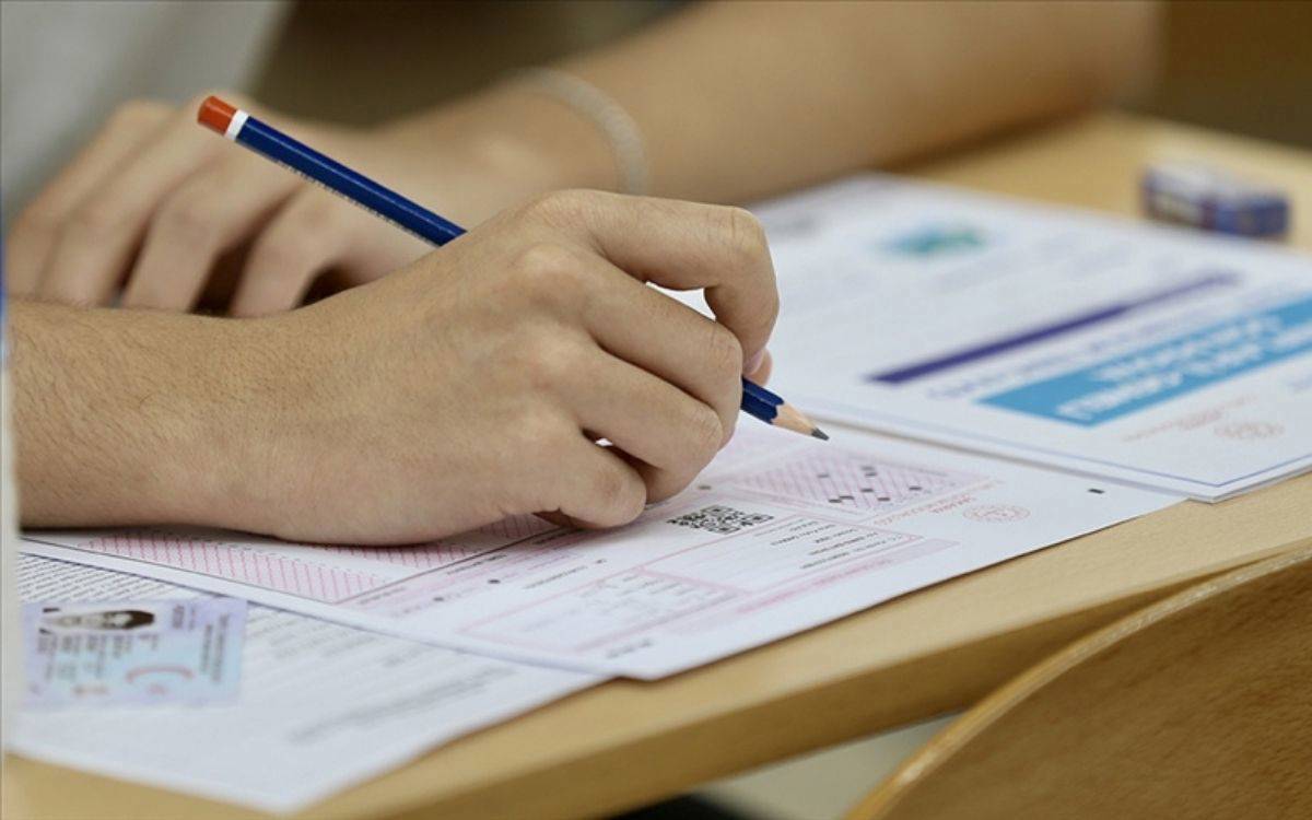 Fotoğrafta bir kişinin sınav salonunda test çözmekte olduğu görülüyor. Kişi elinde mavi-kırmızı uçlu klasik bir kurşun kalem tutuyor ve optik cevap kağıdına işaretleme yapıyor. Masanın üzerinde sınav kitapçığı, optik form ve bir kimlik kartı bulunuyor. Kimlik kartı masanın sol köşesine yerleştirilmiş. Arka planda ahşap bir sıra ve üzerinde silgi seçilebiliyor. Görüntü, sınav ortamının ciddiyetini ve dikkatle soru çözme anını yansıtıyor.