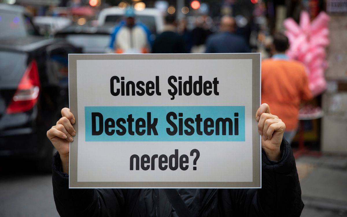 Fotoğrafta kalabalık bir sokakta, bir kişinin elleriyle tuttuğu bir pankart görülüyor. Pankartın üzerinde büyük harflerle “Cinsel Şiddet Destek Sistemi nerede?” yazıyor. Yazının “Destek Sistemi” kısmı açık mavi bir zemin üzerine siyah harflerle vurgulanmış. Arka planda araçlar ve insanlar bulanık şekilde görünüyor; pankart öne çıkarıldığı için dikkat tamamen mesaja odaklanıyor. Görsel, cinsel şiddet mağdurları için yeterli destek mekanizmalarının eksikliğine dikkat çeken bir protesto ya da farkındalık eylemi havası taşıyor.