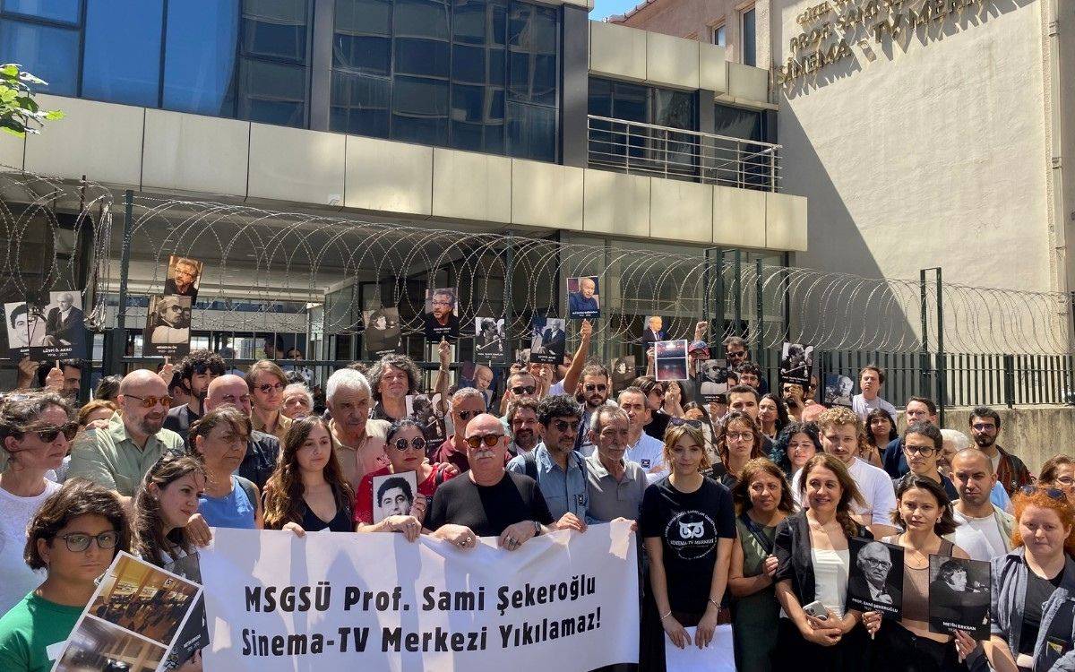 Fotoğraf, kalabalık bir grup insanın bir araya gelerek bir protesto ya da basın açıklaması yaptığını gösteriyor. İnsanlar, bir binanın önünde toplanmış. Binanın duvarında “Prof. Sami Şekeroğlu Sinema-TV Merkezi” yazısı yer alıyor.  Önde duran grup, ellerinde büyük bir pankart tutuyor. Pankartta şu yazıyor: “MSGSÜ Prof. Sami Şekeroğlu Sinema-TV Merkezi Yıkılamaz!”  Katılımcıların bir kısmı ellerinde siyah-beyaz portre fotoğrafları taşıyor. Bu portreler, muhtemelen sinema ve sanat dünyasından önemli isimlere ait. Kalabalık oldukça çeşitli; gençler, kadınlar ve yaşlılar hep birlikte aynı amaçla toplanmış görünüyor. Arkada dikenli tellerin olduğu bir güvenlik bariyeri var, bu da olayın resmi bir kurum ya da üniversite binası önünde gerçekleştiğini düşündürüyor.