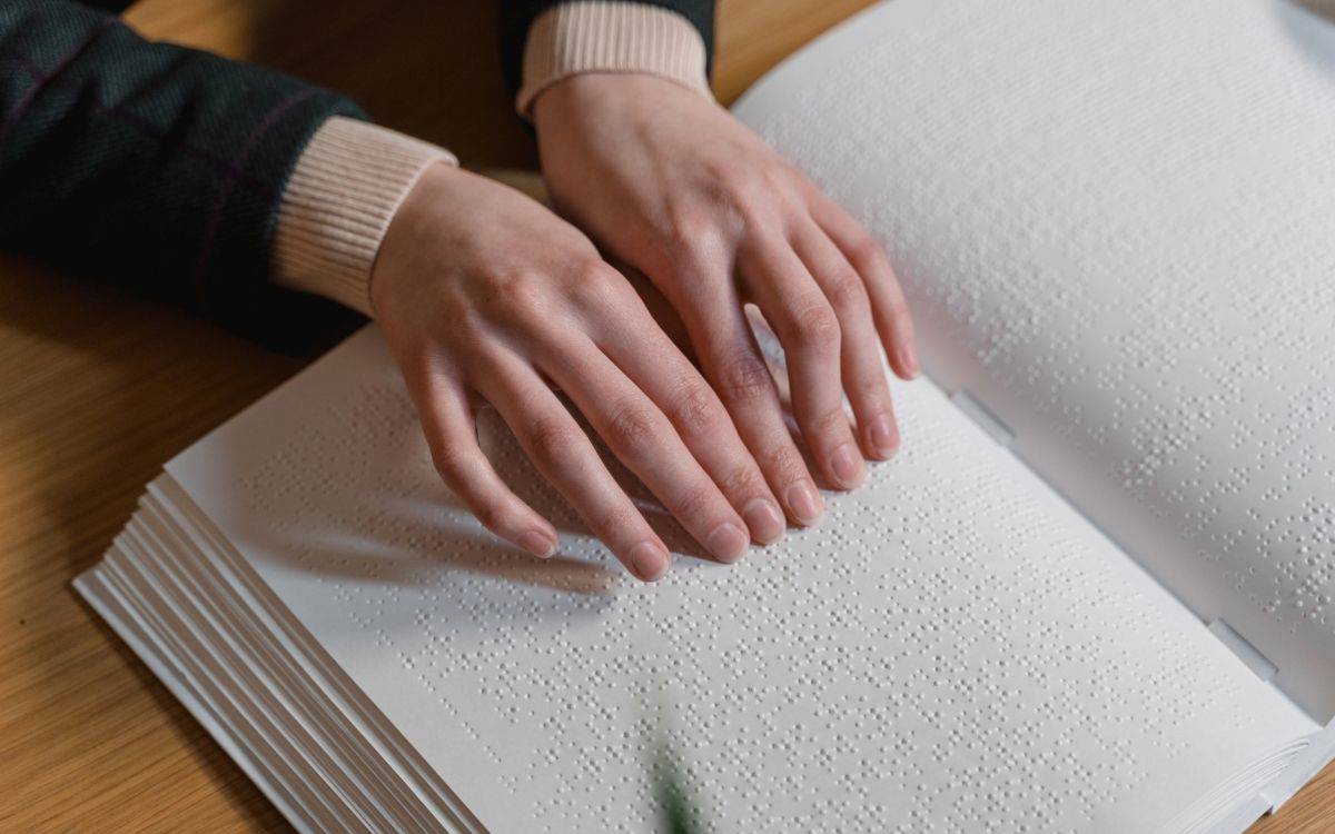 Fotoğrafta, bir kişinin iki eliyle kabartma yazılı (Braille alfabesiyle hazırlanmış) bir kitabı okuduğu görülüyor. Kitap beyaz sayfalardan oluşuyor ve üzerinde kabartmalı noktalar sıralanmış. Kişinin parmakları sayfaların üzerinde geziniyor, yazıyı dokunarak takip ediyor. Üzerinde açık renk manşetli koyu renk bir kıyafet var. Fotoğraf, görme engelliler için hazırlanan Braille kitapların kullanımını ve dokunarak okuma sürecini gösteriyor.