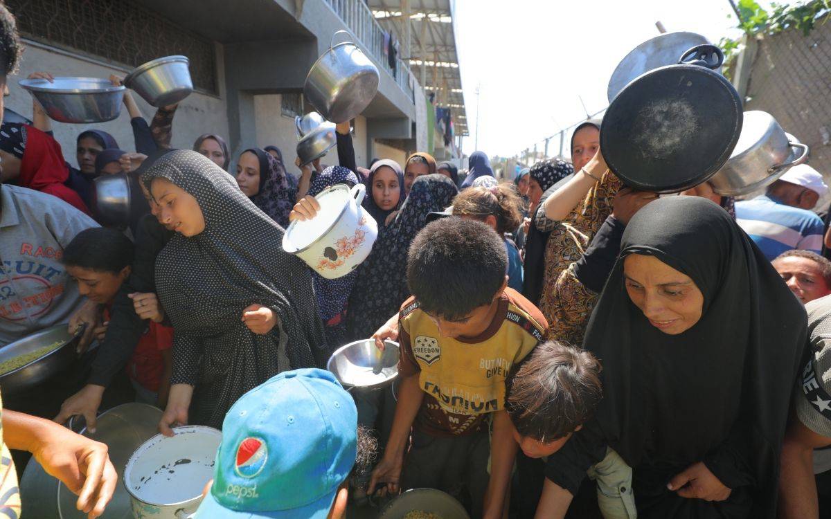 Fotoğrafta kalabalık bir grup insan, açık havada yemek dağıtımında sıraya girmiş görünüyor. Çoğunluğu kadın ve çocuklardan oluşan topluluk, ellerinde farklı boyutlarda tencereler, tabaklar ve kaplarla yemek almayı bekliyor. Ön planda siyah başörtülü bir kadın, yanında ise yemek kabını tutan küçük bir çocuk dikkat çekiyor. Arkada ise kadınların çoğu başörtülü ve yüzlerinde hem yorgunluk hem de umutla bekleyişin izleri var. Bazıları kaplarını havaya kaldırarak yemek almak için dikkat çekmeye çalışıyor.