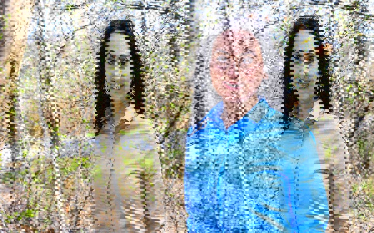 Fotoğrafta, doğada çekilmiş bir portre görülüyor. Arka planda yaprak açmış ağaçlar ve dallar var, hava açık ve aydınlık görünüyor. Ön planda ise açık mavi bir gömlek giymiş, uzun koyu saçlı, gülümseyen bir kadın yer alıyor. Kadın, kulaklarında büyük desenli küpeler takıyor ve doğrudan kameraya bakıyor. Ortam ilkbahar mevsimini andırıyor.