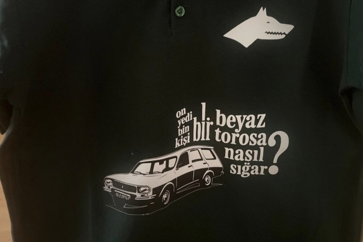 Fotoğrafta koyu renkli bir tişört veya polo yaka bir üst görülüyor. Üzerinde beyaz renkle yapılmış bir tasarım var. Sağ üstte, stilize edilmiş bir kurt başı simgesi yer alıyor. Göğüs kısmında ise eski model bir Renault Toros otomobilin çizimi bulunuyor. Arabanın hemen yanında büyük ve küçük harflerin karışık biçimde yer aldığı bir yazı dikkat çekiyor: “On yedi bin kişi bir beyaz torosa nasıl sığar?” Bu ifade, hem görsel olarak hem içerik olarak dikkat çekici biçimde tasarlanmış. Metin, Türkiye’nin 1990’lı yıllarındaki faili meçhul cinayetlerle özdeşleşmiş “beyaz Toros” kavramına gönderme yapıyor. Arabanın altındaki plaka da okunaklı biçimde çizilmiş. Arka plan siyah ya da koyu yeşil tonlarında; tüm baskı detayları beyaz renkte. (Görsel yapay zeka yardımıyla betimlenmiştir)