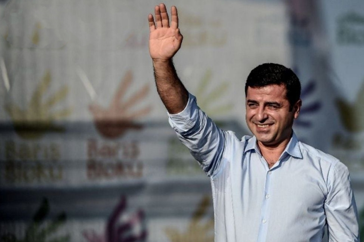 Fotoğrafta, Selahattim Demirtaş açık mavi gömlek giymiş olarak görülüyor. Gülümsüyor ve sağ elini havaya kaldırarak selam verir gibi duruyor. Arka planda, üzerinde çok sayıda renkli el sembolü bulunan bir afiş veya pankart var; afişte “Barış Bloku” yazısı tekrarlanıyor. Görsel genel olarak açık havada, gündüz çekilmiş gibi görünüyor. Işık figürün yüzüne yandan vuruyor ve sahne atmosferi sıcak, pozitif bir his taşıyor. (Görsel yapay zeka yardımıyla betimlenmiştir)