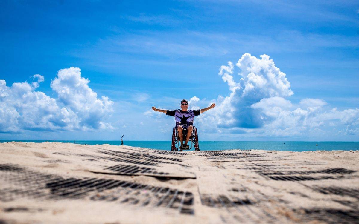 Fotoğrafta tekerlekli sandalyede oturan bir kişi, kollarını iki yana açmış şekilde deniz kıyısında poz veriyor. Kişinin arkasında uçsuz bucaksız mavi bir deniz ve gökyüzünü kaplayan beyaz bulutlar görülüyor.