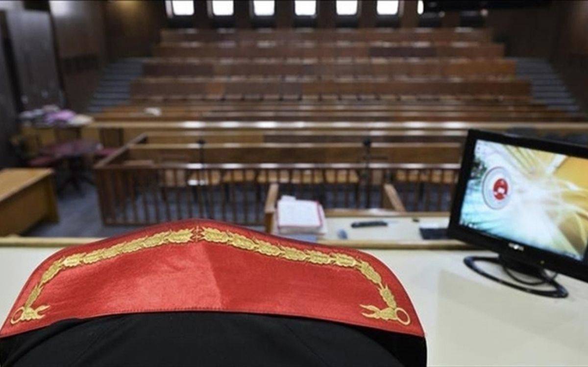 Fotoğrafta bir mahkeme salonu görülüyor. Ön planda, hâkimlerin cübbelerinde yer alan kırmızı kadife kumaş üzerine altın sarısı işlemeli desen dikkat çekiyor. Bu detay, hâkim kürsüsünün hemen önünde, yargı otoritesini temsil eden bir konumda yer alıyor. (Bu betimleme yapay zeka ile yapıldı)