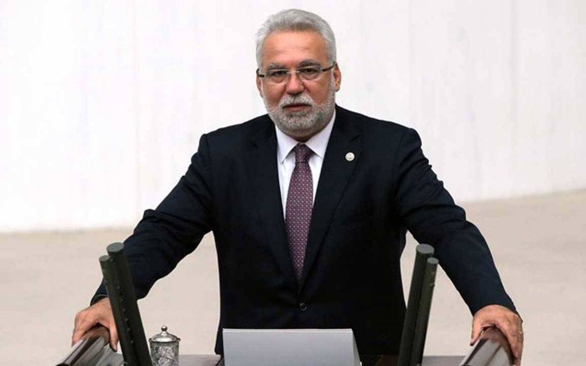 Fotoğrafta takım elbiseli, beyaz saçlı ve sakallı bir erkek, resmi bir ortamda — muhtemelen bir meclis kürsüsünde — ayakta duruyor. Üzerinde koyu renk bir takım elbise, beyaz gömlek ve bordo bir kravat bulunuyor. ( Bu betimleme yapay zeka ile yapıldı)