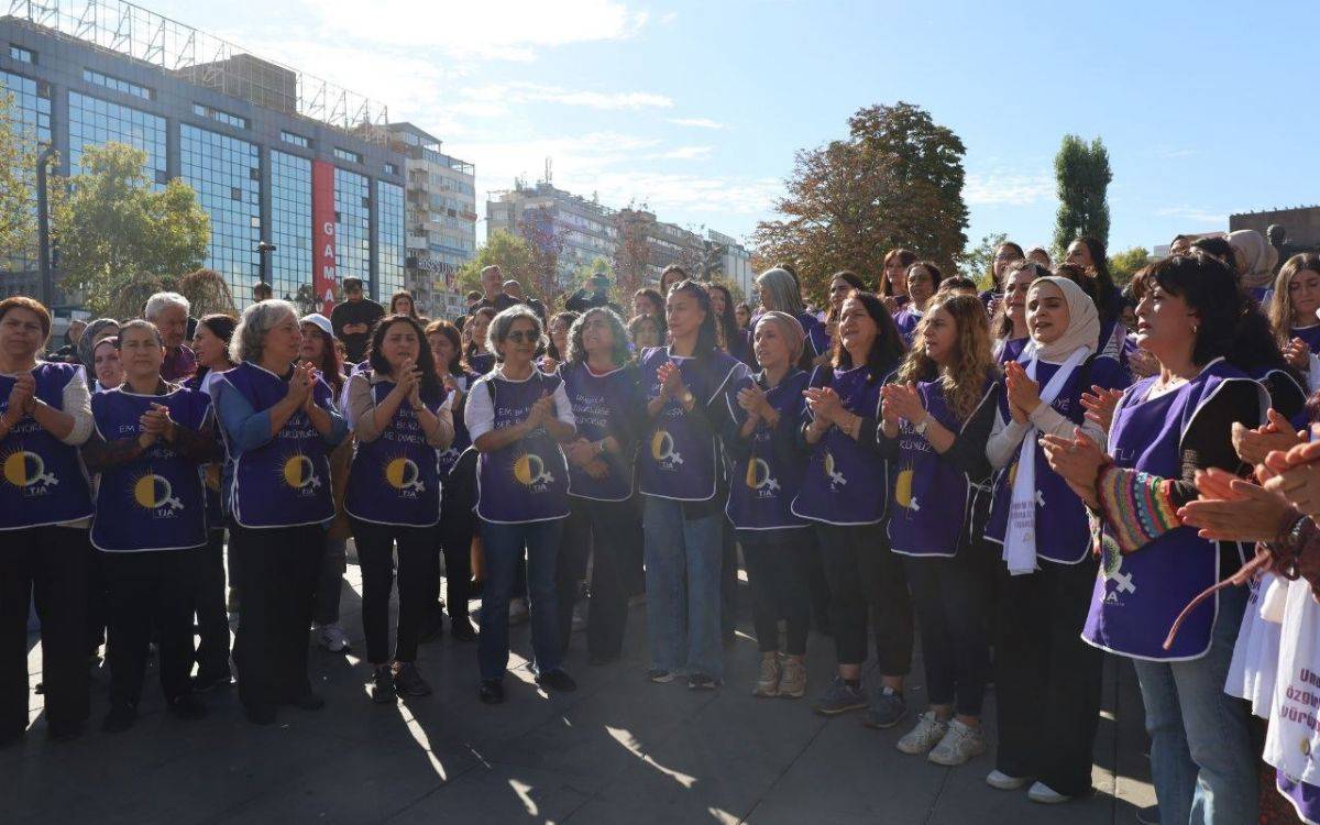 Fotoğrafta açık havada toplanmış bir grup kadın görülüyor. Kadınların çoğu mor renkli, üzerinde sarı güneş ve beyaz güvercin sembolleri bulunan yelekler giymiş. Yeleklerde “TJA” kısaltması yer alıyor. Kadınlar halka şeklinde durarak alkış tutuyor ve birlikte slogan atıyor ya da şarkı söylüyor gibi görünüyorlar.