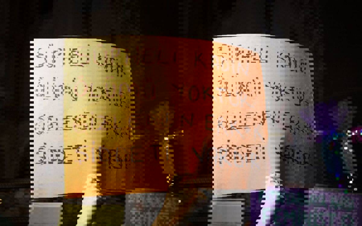 Fotoğrafta, bir protesto veya yürüyüş sırasında çekilmiş bir sahne yer alıyor. Görselde bir kişi elinde turuncu bir karton pankart tutuyor. Pankartın üzerinde siyah kalemle büyük harflerle şu cümle yazılı: “ŞÜPHELİ KADIN ÖLÜMÜ YOKTUR. GİZLENEN ERK*K ŞİDDETİ VARDIR.” Bu görseli yapay zeka betimledi.