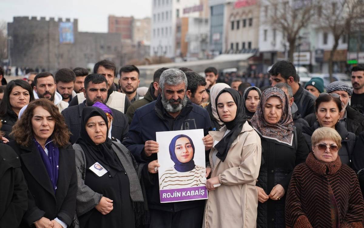 Fotoğrafta, bir grup insanın Rojin Kabaş adlı genç bir kadının portresini taşıdığı bir anma veya protesto etkinliği görülüyor. Kalabalık, kent merkezinde – muhtemelen Diyarbakır’da, çünkü arka planda şehrin ünlü surları seçiliyor – toplanmış durumda.