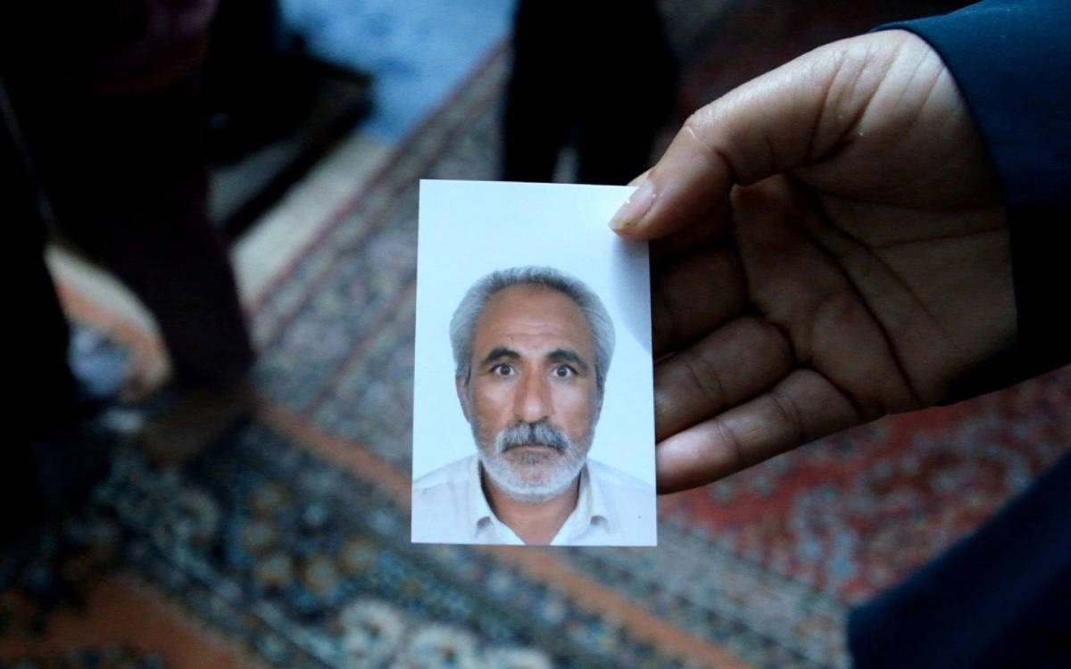 Fotoğrafta bir el, üzerinde orta yaşlı bir erkeğin portre fotoğrafı bulunan küçük bir vesikalık fotoğrafı tutuyor. Erkek, gri saçlı ve sakallı, beyaz gömlek giymiş. ( yapay zeka ile betimlendi)