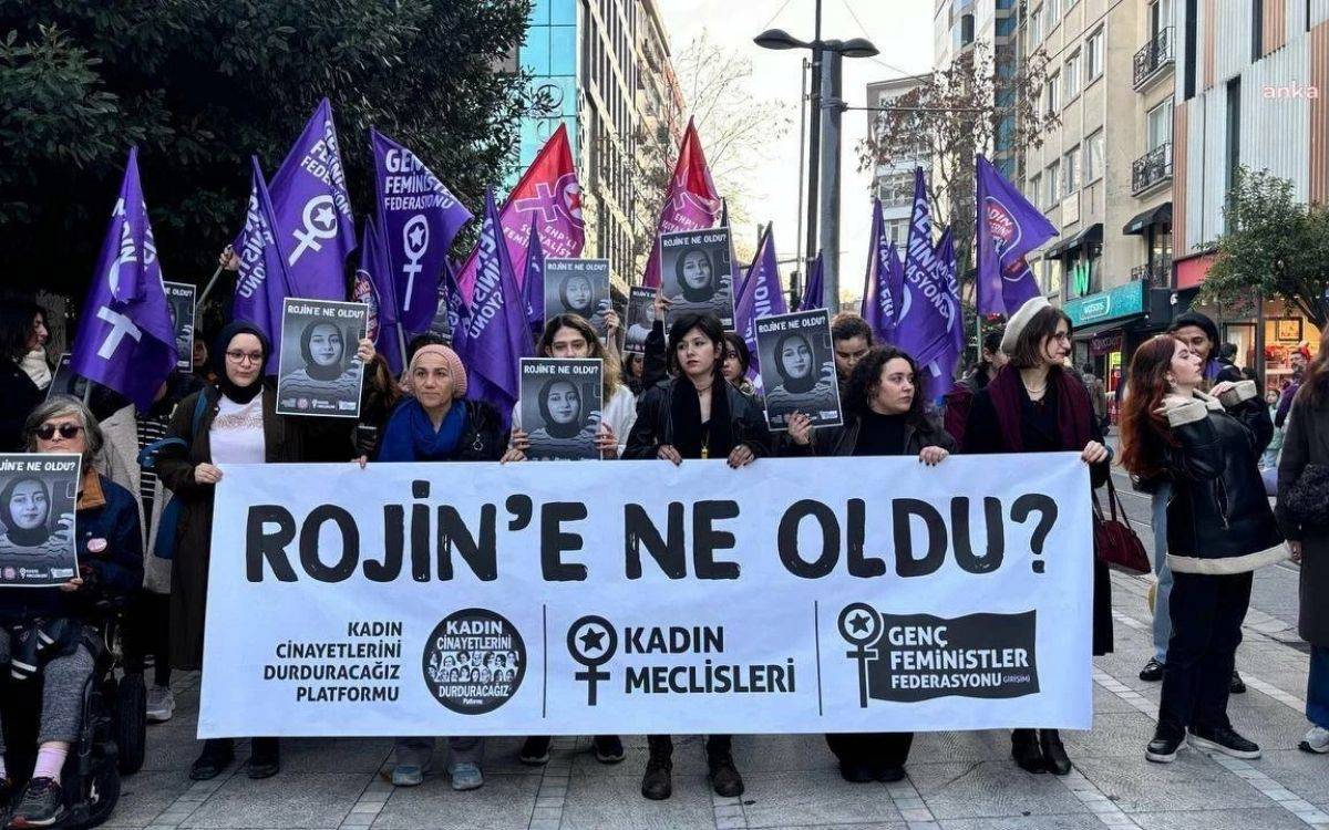 Fotoğrafta bir grup kadın, şehir merkezinde düzenlenen bir protestoda görülüyor. Grup, ellerinde “Rojin’e ne oldu?” yazılı büyük bir pankart taşıyor. Pankartın altında “Kadın Cinayetlerini Durduracağız Platformu”, “Kadın Meclisleri” ve “Genç Feministler Federasyonu” logoları yer alıyor.