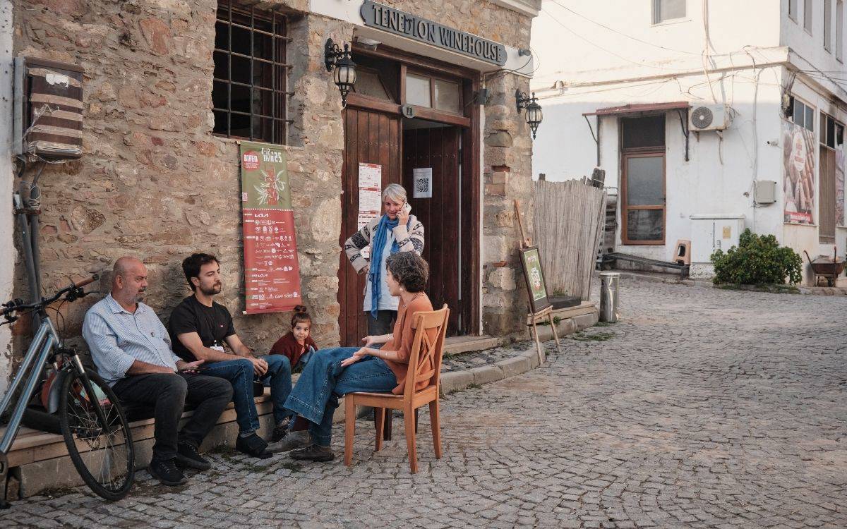 Görselde taş duvarlı, eski bir binanın önünde bir grup insan oturmuş sohbet ediyor. Binanın kapısında “Tenetlon Winehouse” yazılı bir tabela bulunuyor. Kapının önünde, biri telefonla konuşan beş yetişkin ve bir çocuk yer alıyor.