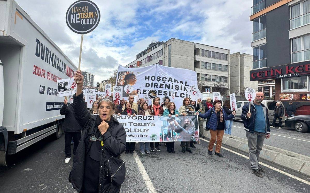 Fotoğrafta bir grup insan, şehir içinde bir caddede yürüyüş yapıyor. Katılımcıların ellerinde dövizler ve pankartlar var. En öndeki pankartta “Hakan Tosun’a ne oldu?” ifadesi yer alıyor.