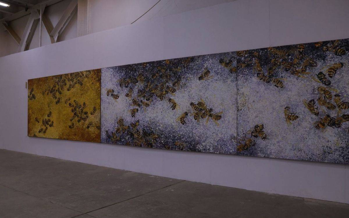 Fotoğrafta bir sergi alanında, geniş bir duvara asılmış büyük boyutlu üç parçadan oluşan (tripti̇k biçiminde) bir tablo yer alıyor. Eserin her bir panelinde onlarca arı figürü görülüyor. Arılar, gerçekçi bir biçimde ama yoğun fırça darbeleriyle betimlenmiş. Sol panel sıcak sarı ve altın tonlarında, ortadaki ve sağdaki paneller ise daha çok mor, beyaz ve gri tonlarıyla boyanmış.