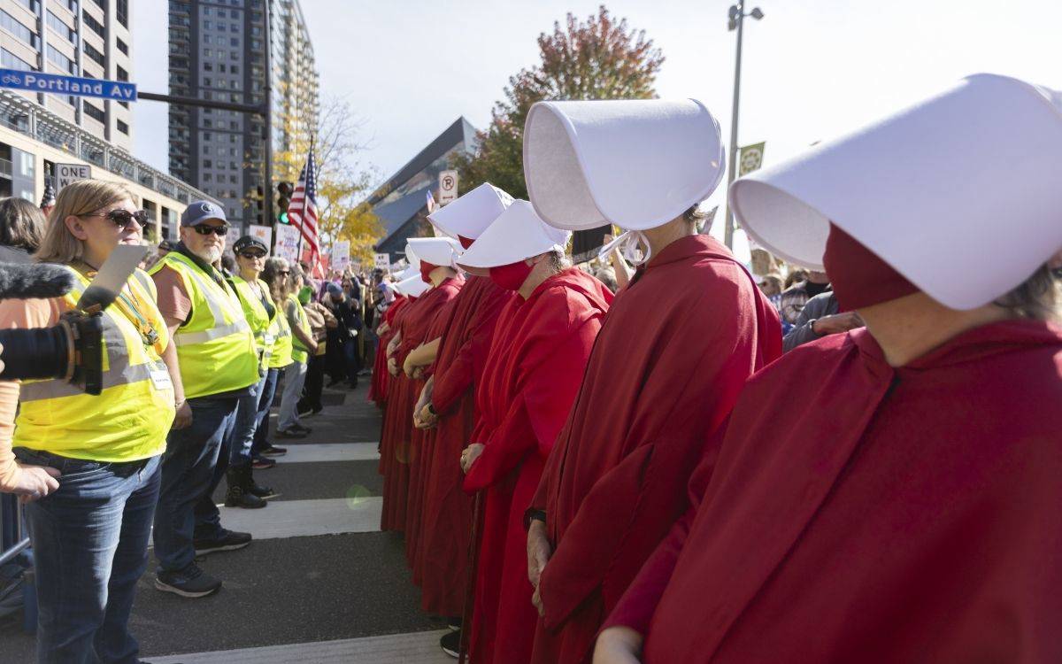 Fotoğrafta, bir protesto gösterisi sırasında iki grup insan karşılıklı olarak duruyor. Sağ tarafta, kırmızı pelerin ve beyaz başlıklar giymiş bir grup